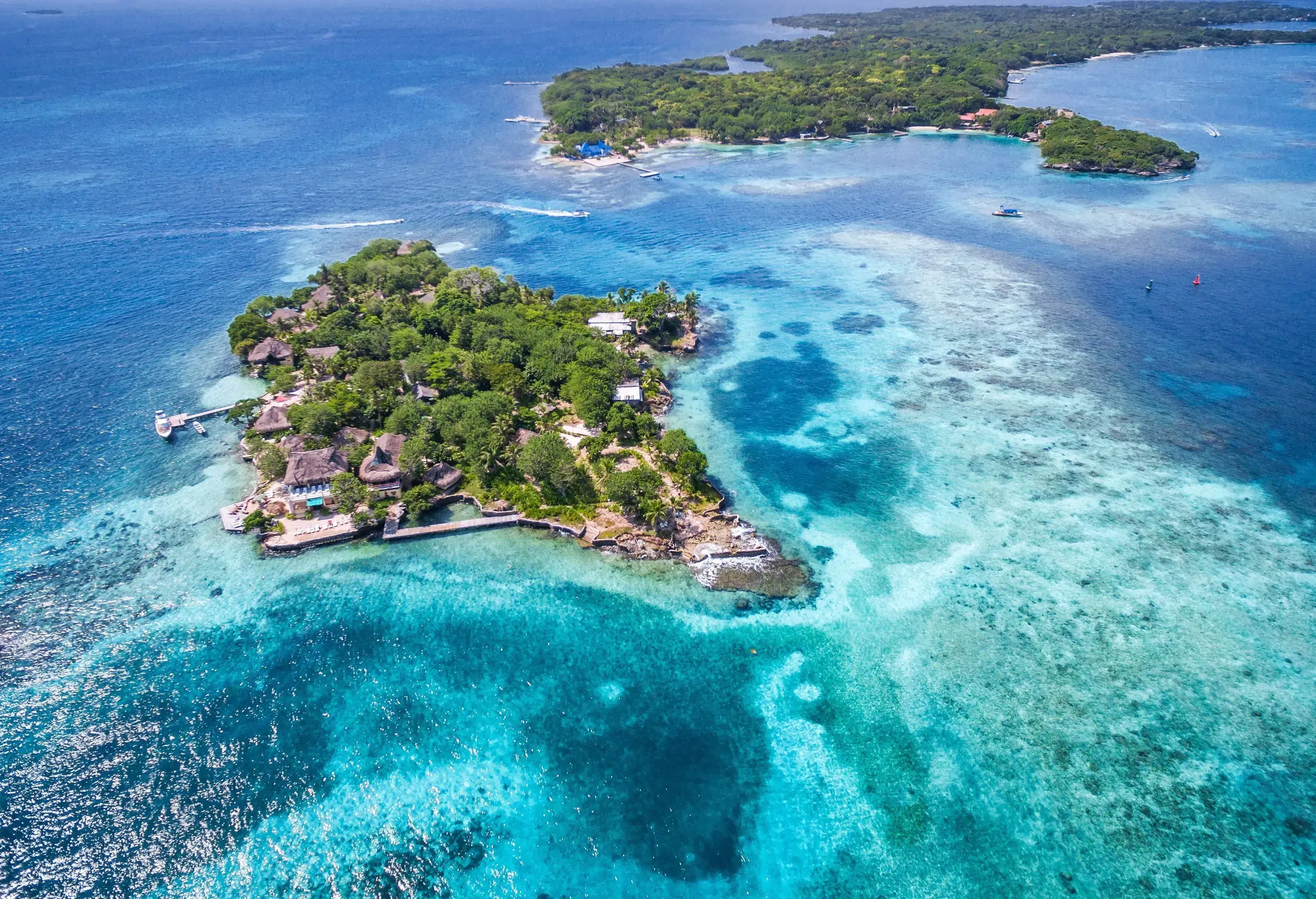 A forested islet with visible thatched houses emerges on the crystal-clear turquoise sea near an elongated island.