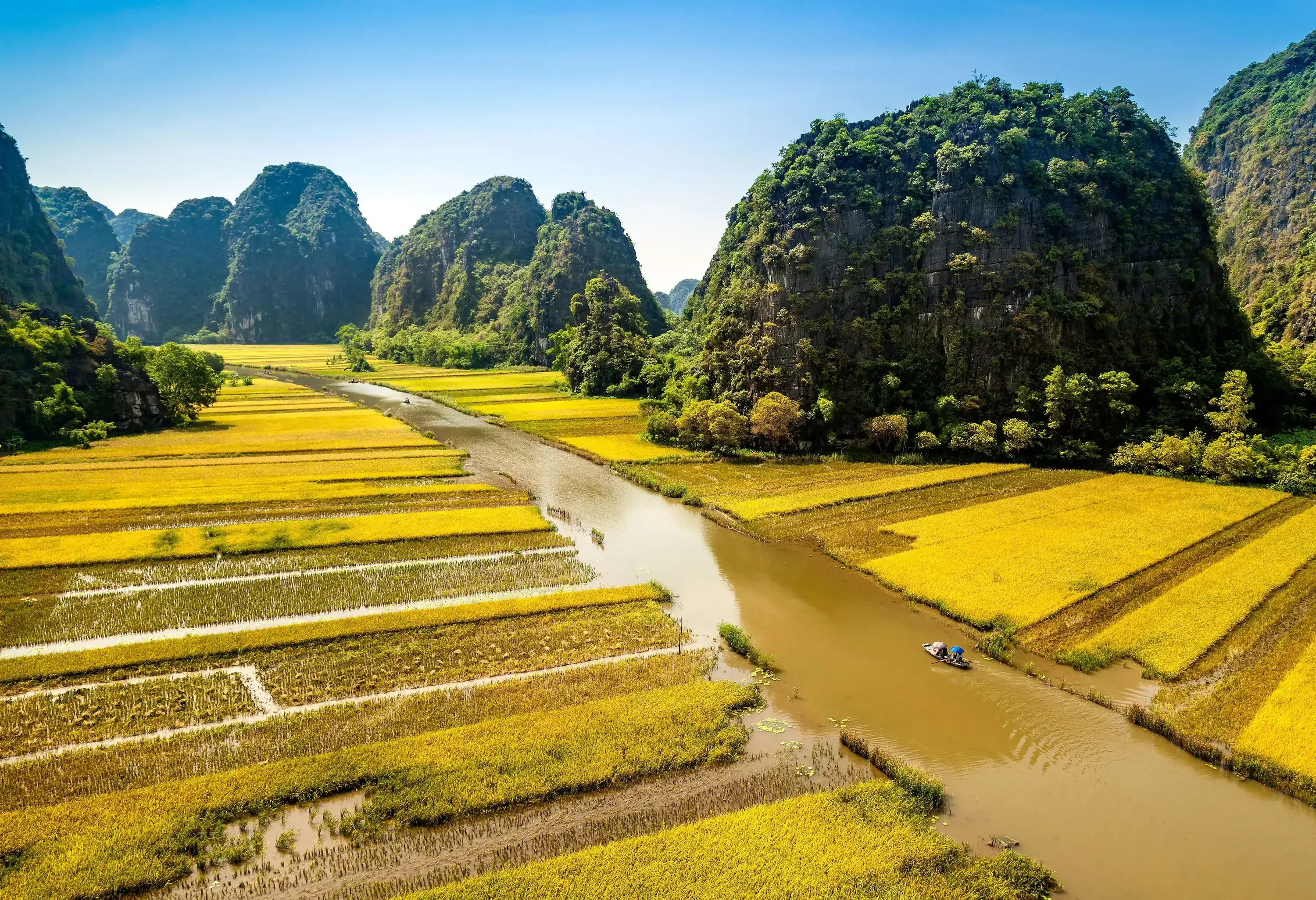 A small boat sails on the muddy river, splitting the rice field into two among the rocky hills.
