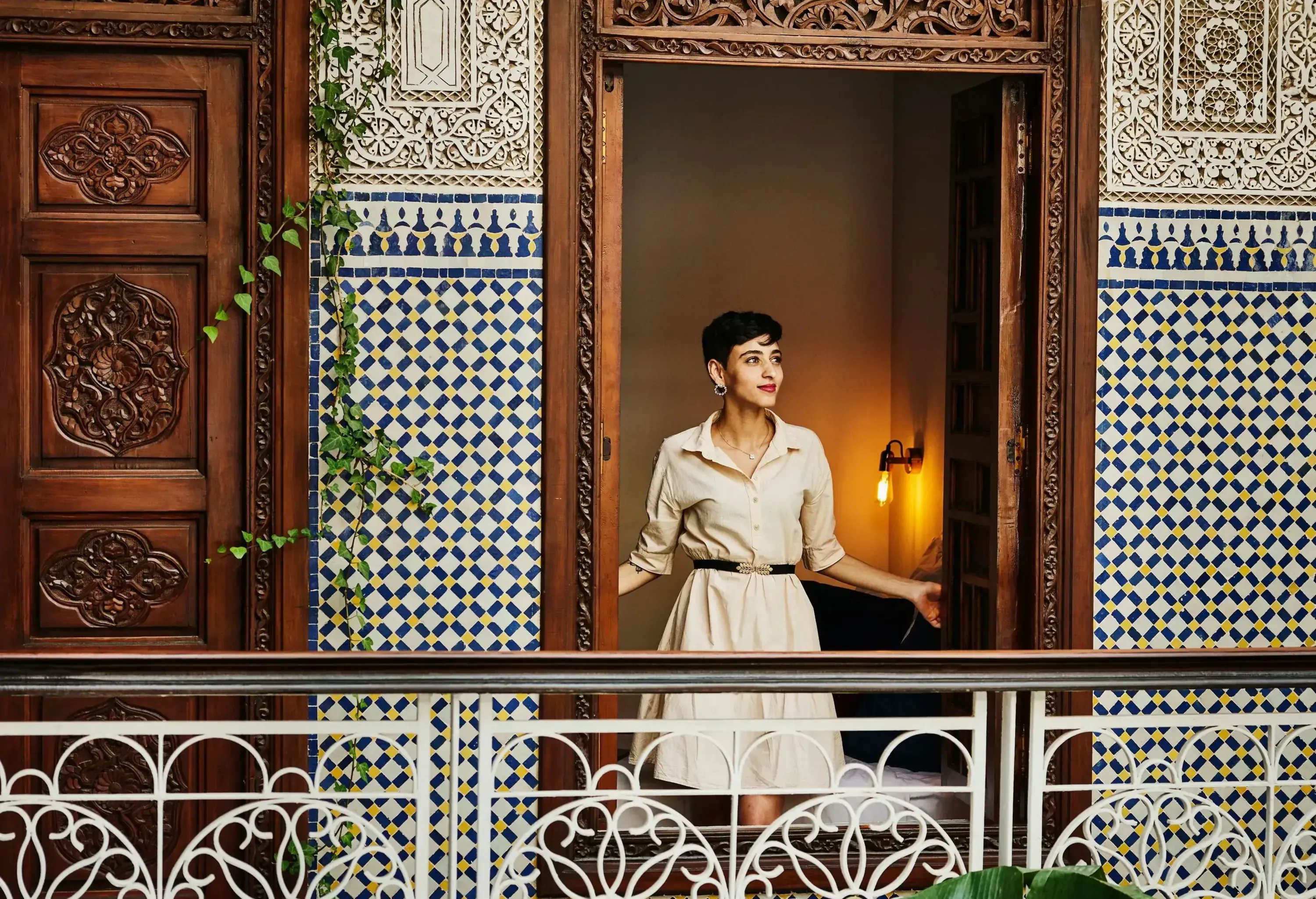 A person stands and looks out from an open ornate wooden doorway on a balcony with decorative tile walls and patterned railings.