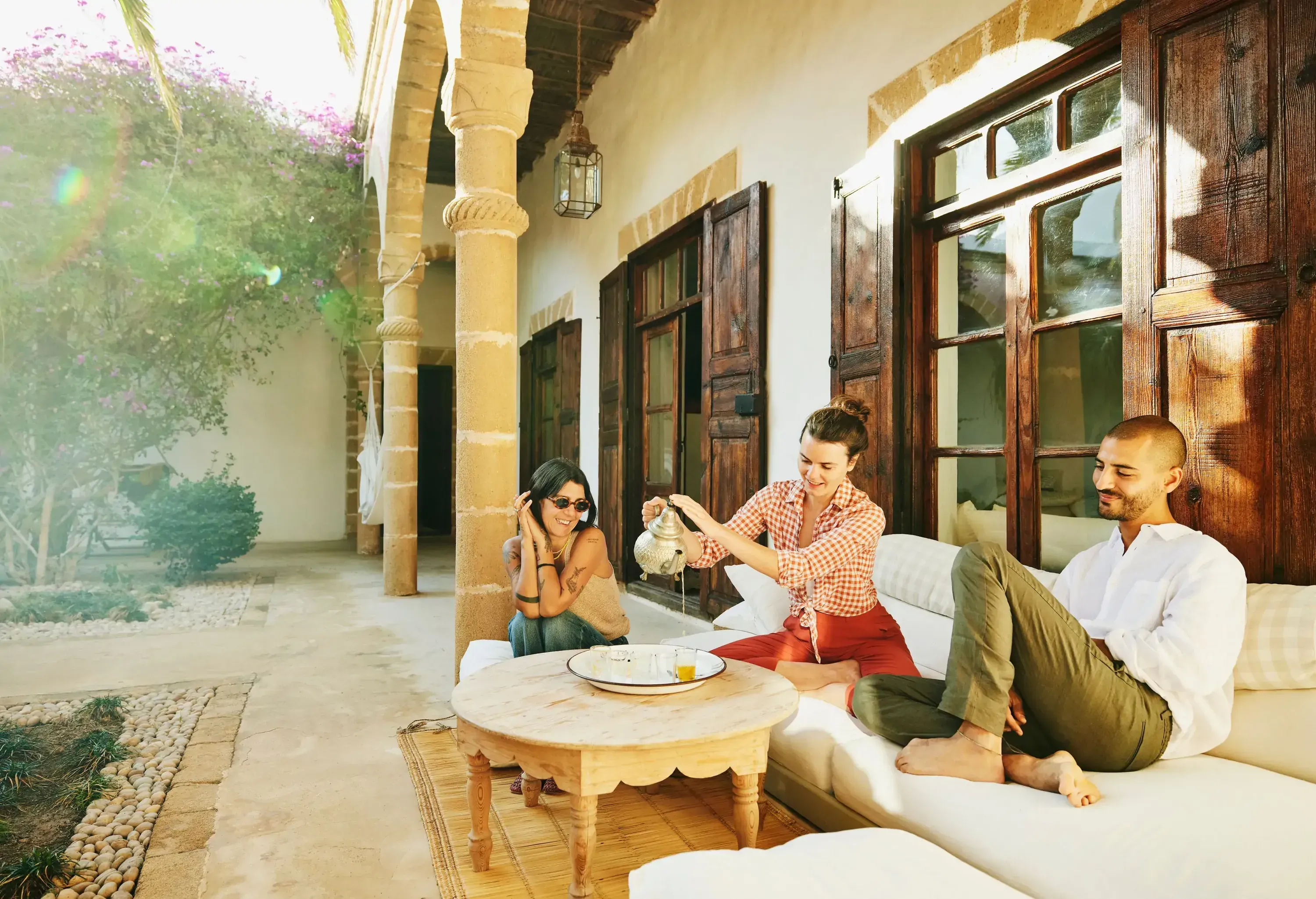 Wide shot of smiling woman pouring afternoon tea for friends while relaxing in courtyard of tropical villa during vacation in Morocco