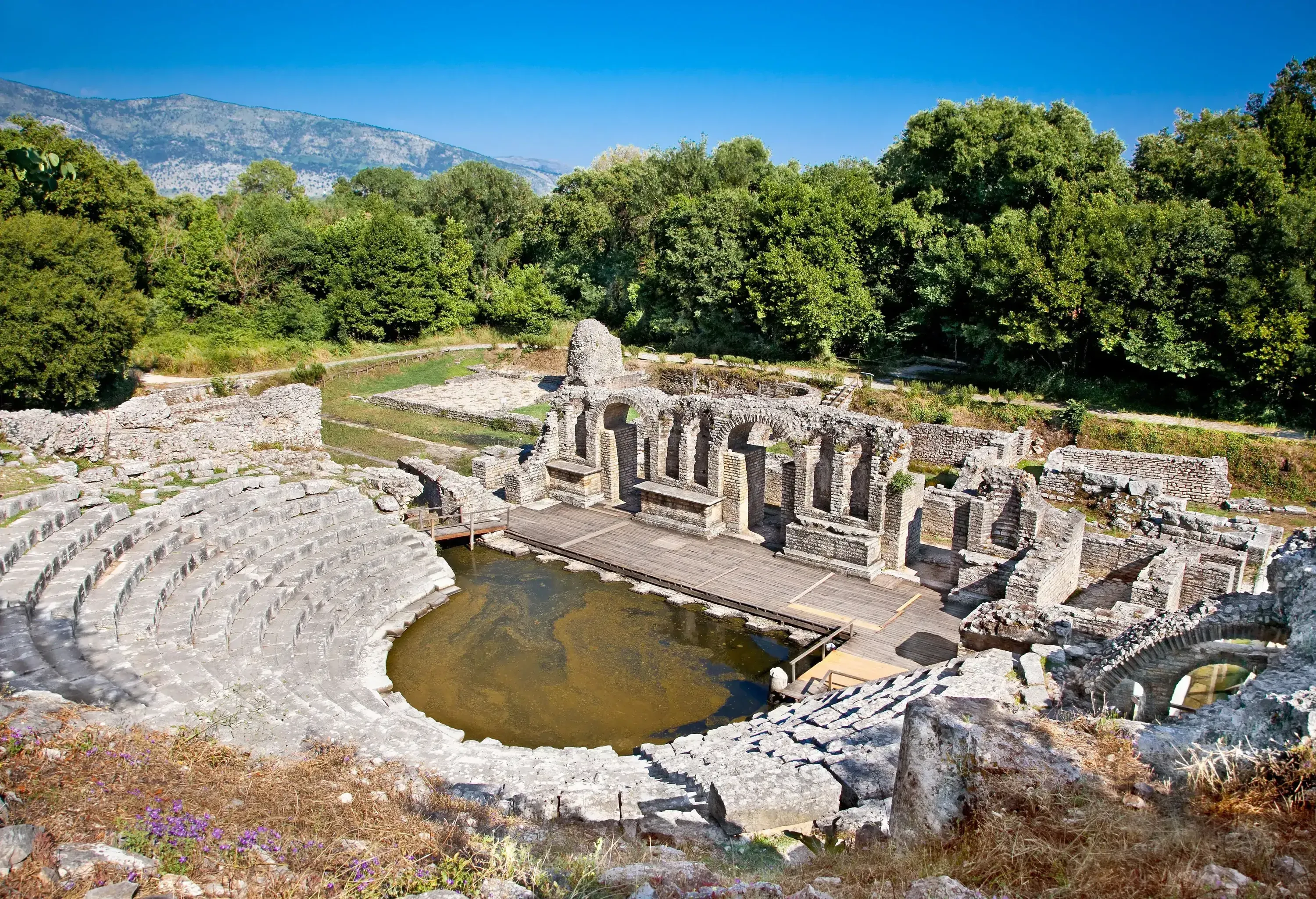 Ruins of an ancient amphitheatre with semi-circular terraced seats and a stage with damaged archways.