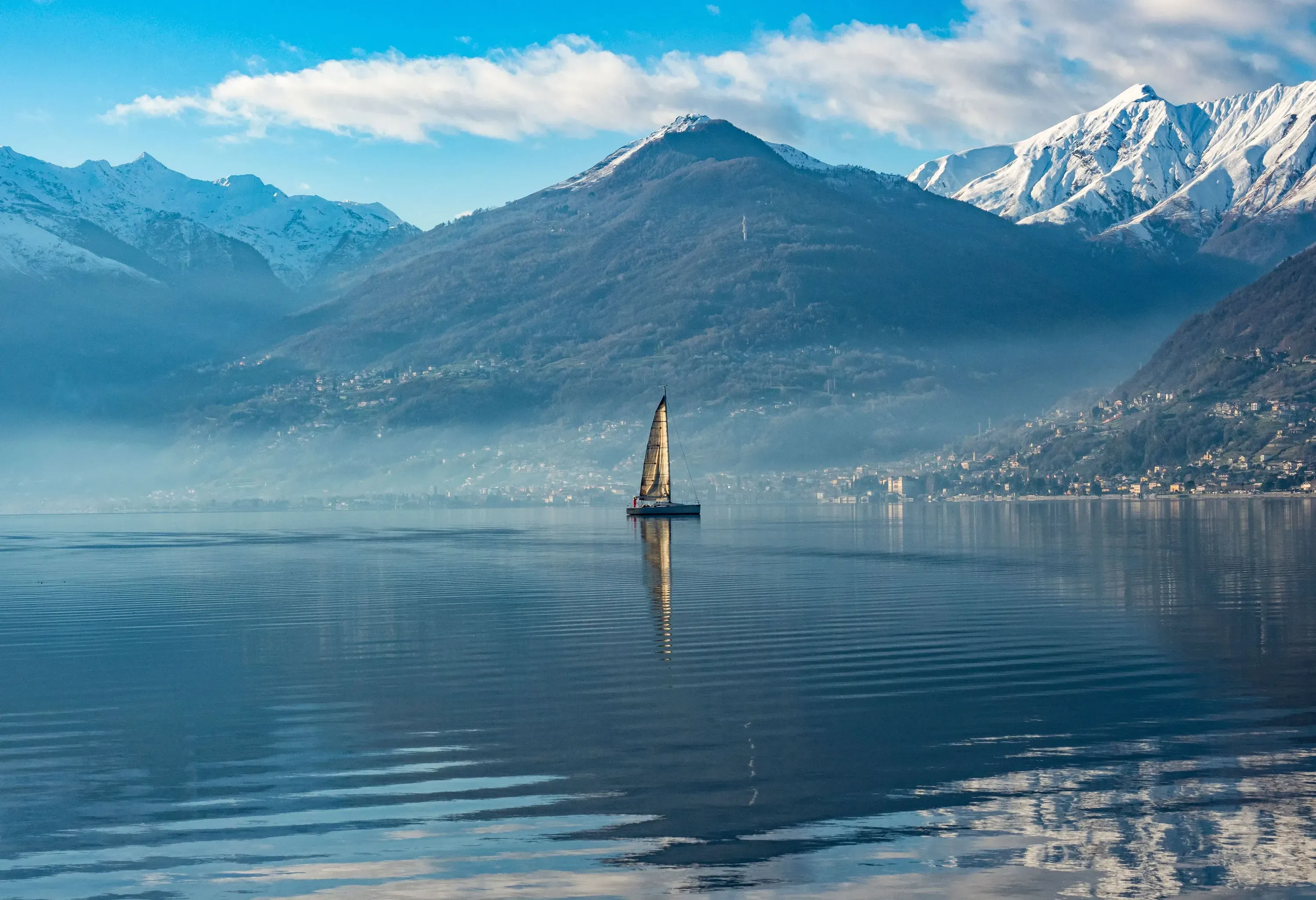 A sailboat glides on a calm, misty lake, with its reflection visible on the water, against a backdrop of mountains.